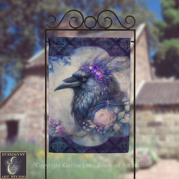 a banner with a large, detailed illustration of a bird, possibly a crow, wearing a headdress with purple and blue elements. The bird is surrounded by flowers and foliage, creating a whimsical and artistic scene. The banner is displayed on a metal stand, and there is a blurred background that includes a building and trees.